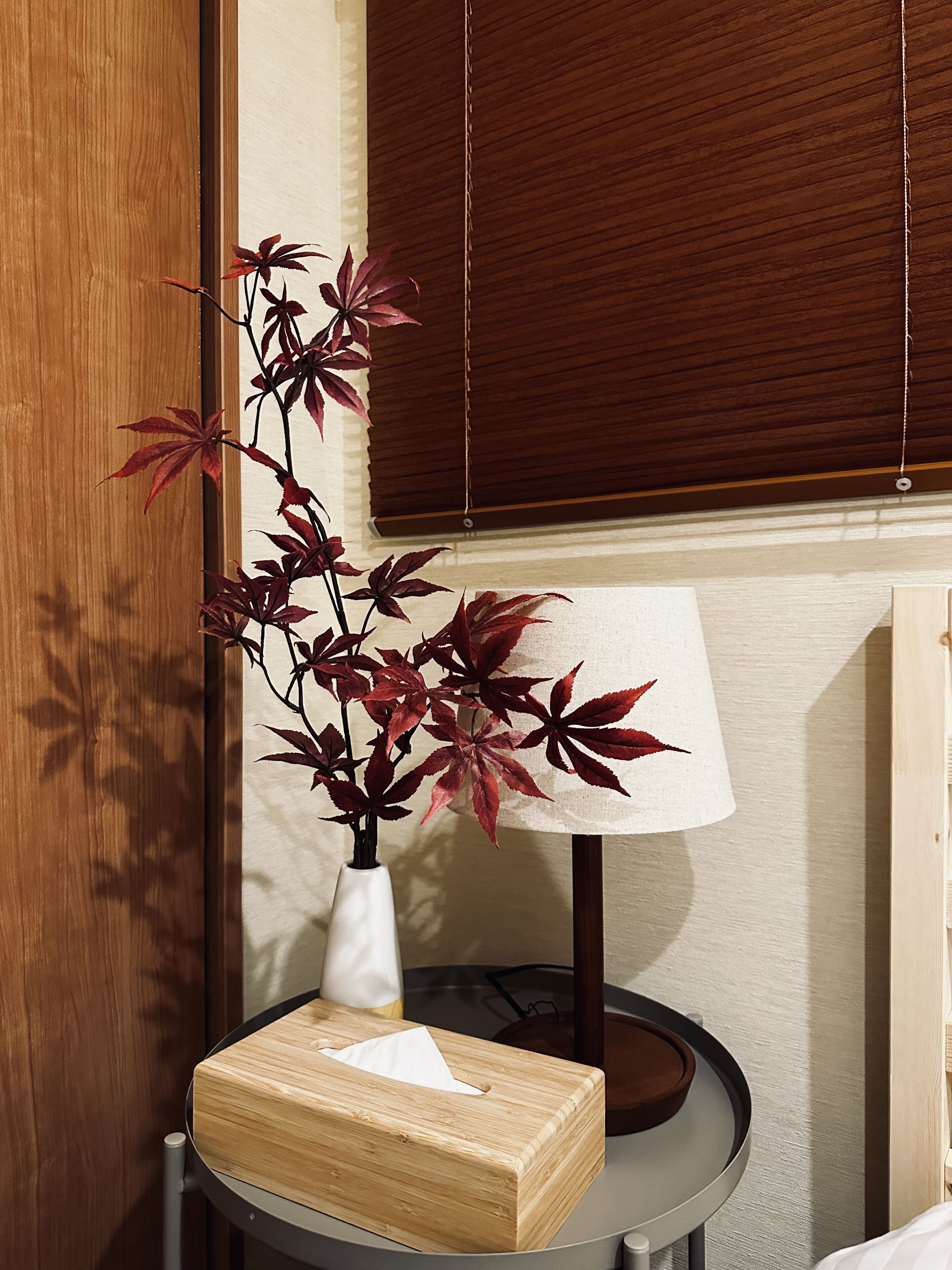 A wooden bedside table with a white vase containing red leaves, a tissue box, and a lamp.
