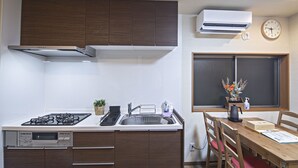 Een kleine keuken met een fornuis, spoelbak en houten kastjes. Er is een raam met uitzicht, een klok en een airconditioner aan de muur.