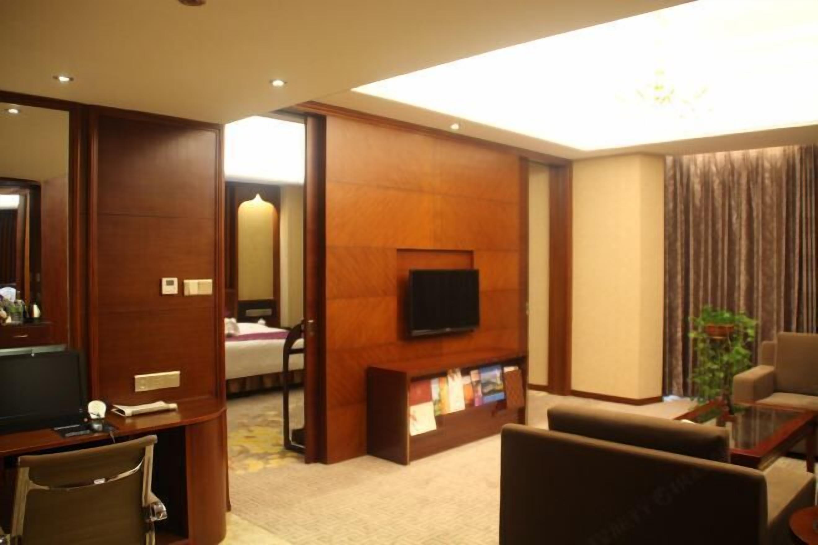 A hotel room with a TV, a desk, a chair, a sofa, a bookshelf, and a mirror.