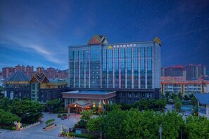 A modern hotel with a glass facade, surrounded by greenery and other buildings. - Guowei Hotel (Menghai)