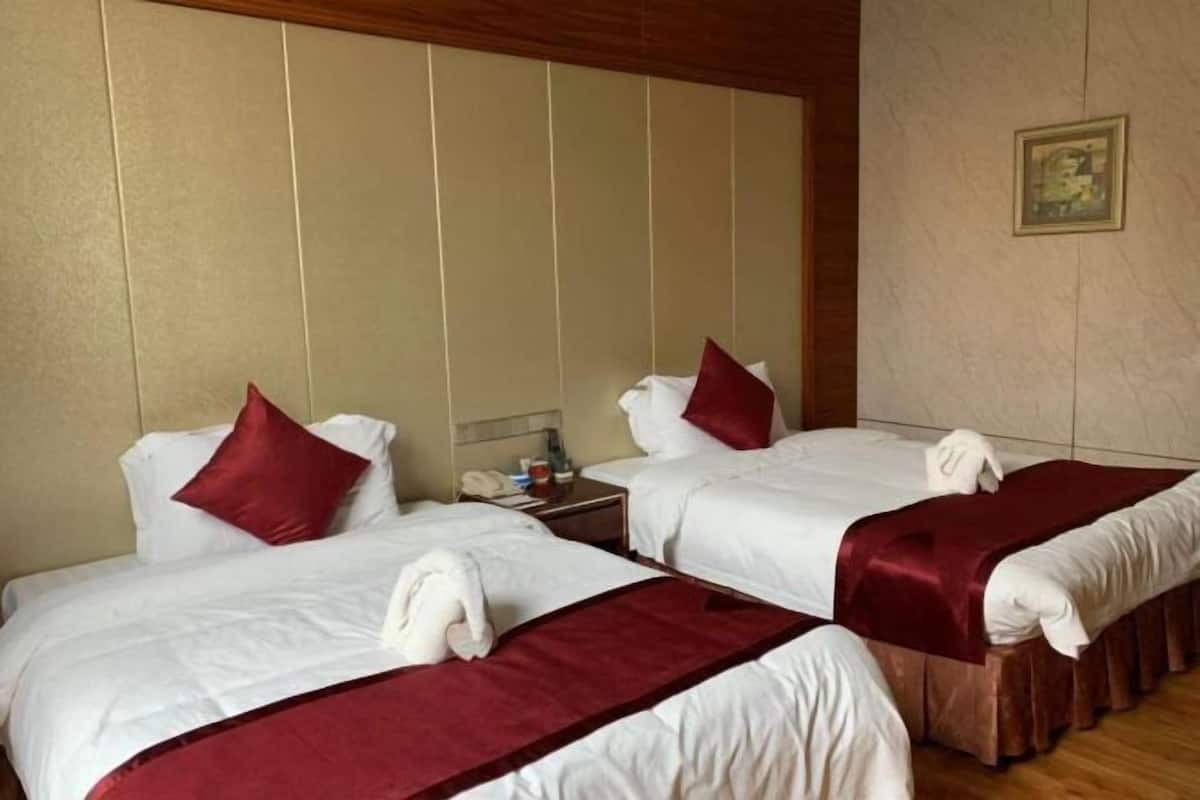 Habitación de hotel con dos camas, cada una con sábanas blancas y almohadas rojas. Cuenta con un cabecero de madera, una mesita de noche con lámpara y un cuadro en la pared.