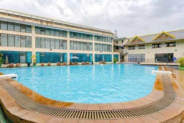 Une piscine avec une terrasse ensoleillĂ©e, un grand bĂątiment dotĂ© de nombreuses fenĂȘtres et un ciel dĂ©gagĂ©.