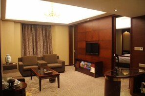 A hotel room with a sofa set, a coffee table, a flat-screen TV mounted on a wooden panel, and a bed with a headboard. - Guowei Hotel (Menghai)