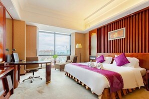 Habitación de hotel con una cama grande, un escritorio, una silla y vistas de la ciudad a través de amplias ventanas.