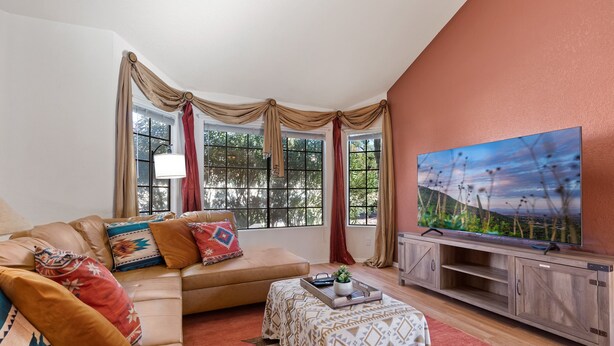 A living room with a beige sofa, colorful cushions, a coffee table with a tray, a TV on a wooden stand, and large windows with curtains. - Southwestern Paradise, Simply Classic (Oro Valley)