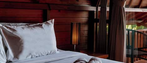 Una cama con sábanas blancas y dos toallas enrolladas, paredes de madera y una ventana con cortinas.