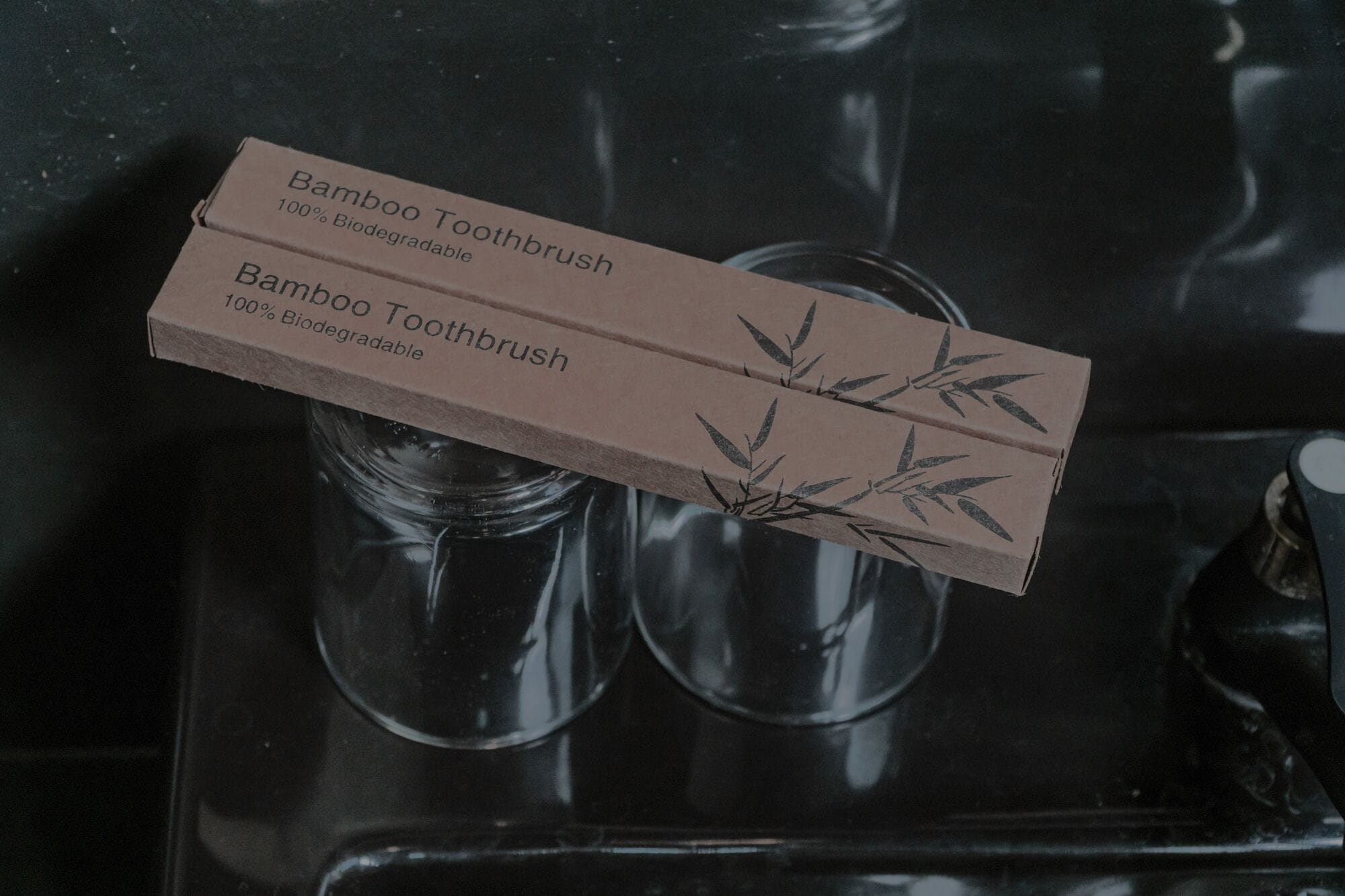 Un estuche de bambú para cepillos de dientes con diseño de hoja de bambú, sobre una superficie oscura junto a dos vasos de cristal.