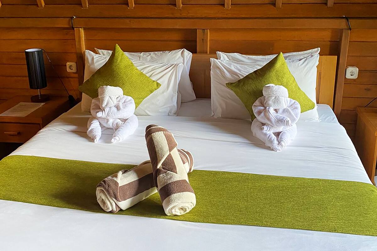 A hotel room with a wooden ceiling, a bed with white linens, two green pillows, and two towels folded on the bed.