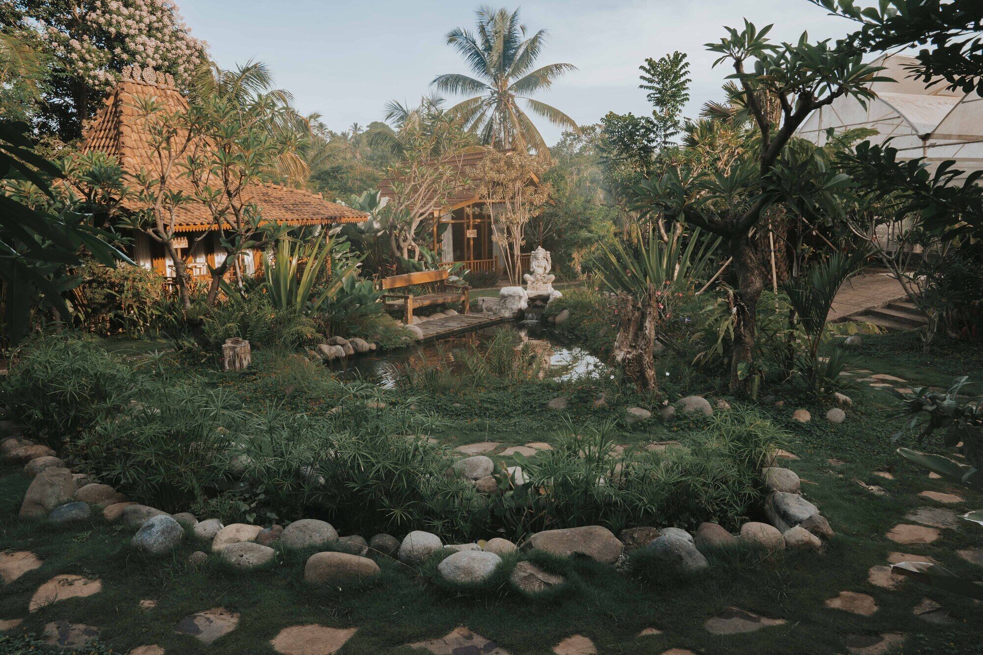 A tropical garden with a pond, stone pathway, and rustic wooden structures.