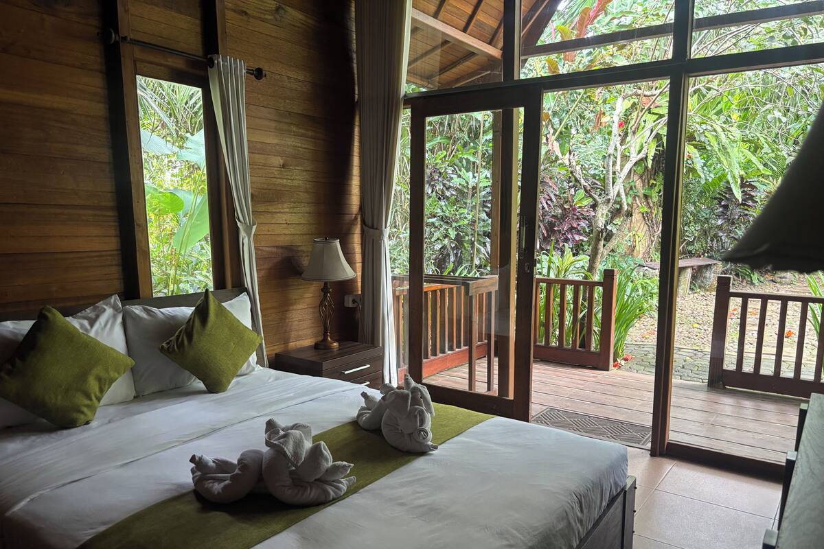 A wooden-framed room with a bed, pillows, a lamp, and a view of a wooden deck and greenery.