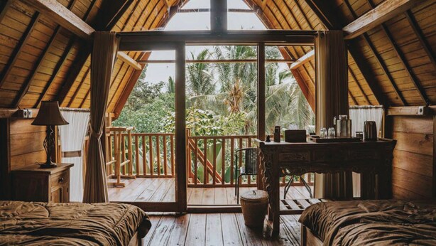 A cozy room with a wooden ceiling, a large sliding glass door leading to a balcony, and a small table with a lamp. - Kusfarm Bali by AHM (Selemadeg)