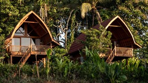 Two wooden cabins with balconies, surrounded by lush tropical vegetation. - Kusfarm Bali by AHM (Selemadeg)