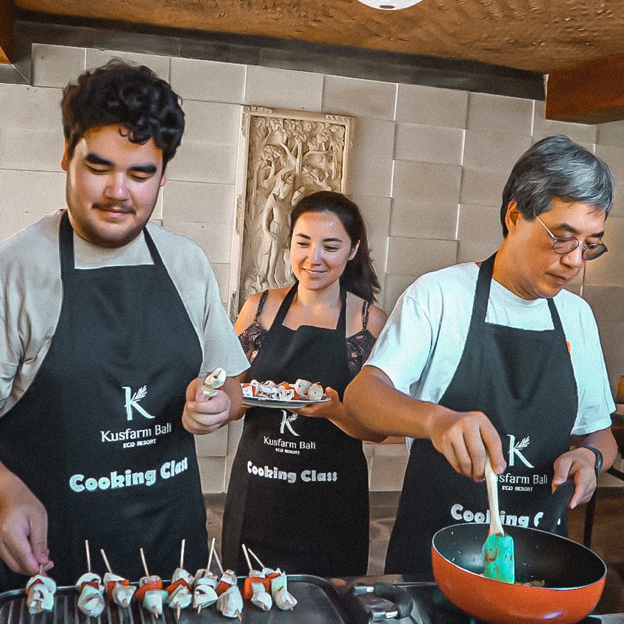 Three individuals wearing aprons with the logo 'Kusfarm Bali' engaged in cooking activities.