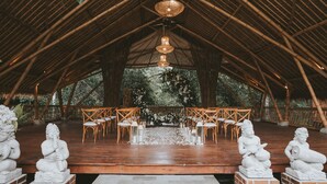 An indoor venue with a high bamboo ceiling, wooden chairs arranged in rows, and white flower petals on the floor. - Kusfarm Bali by AHM (Selemadeg)