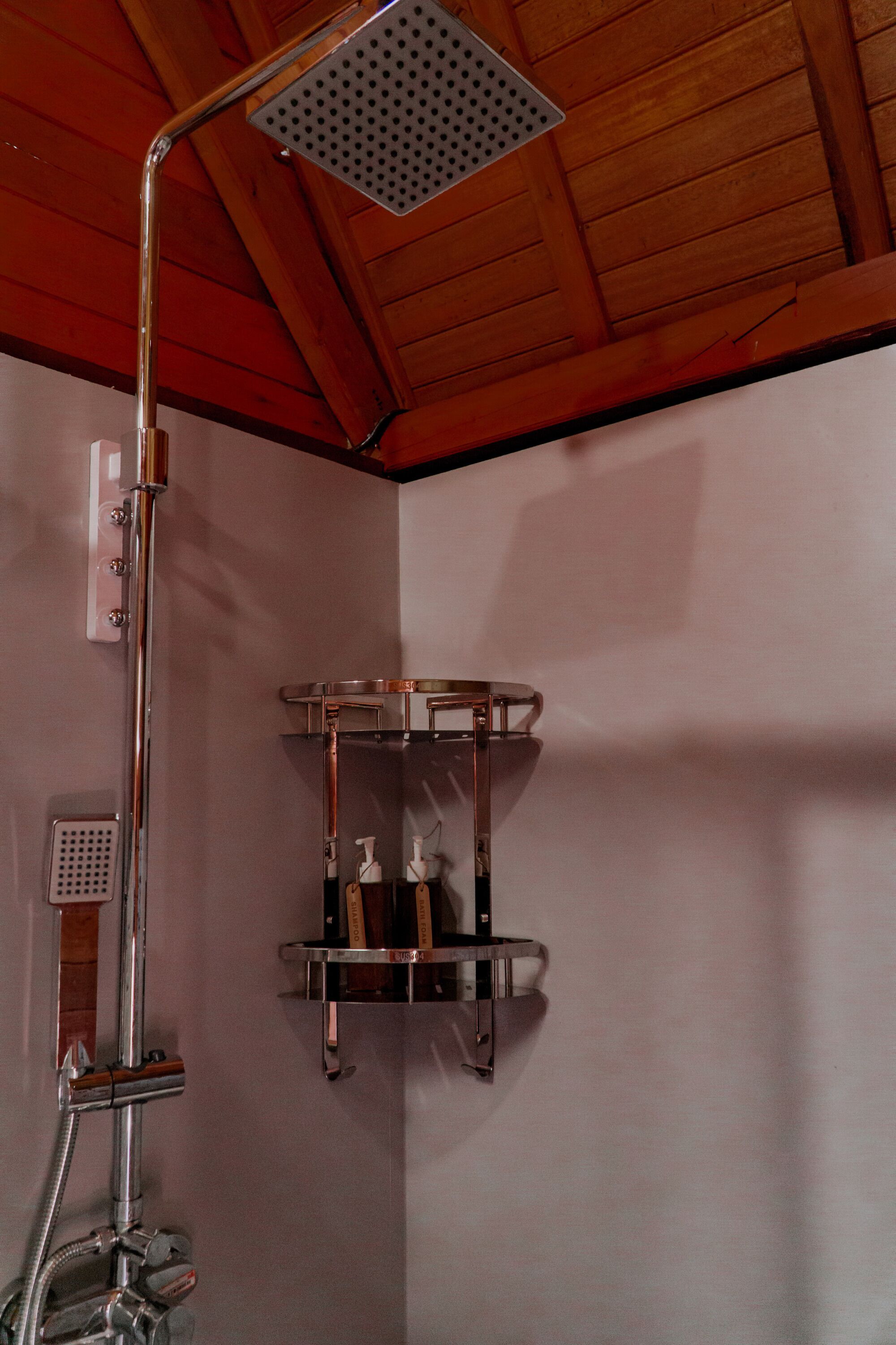 Una ducha con cabezal de ducha de mano y llave de pared. Hay un estante con artículos de aseo.