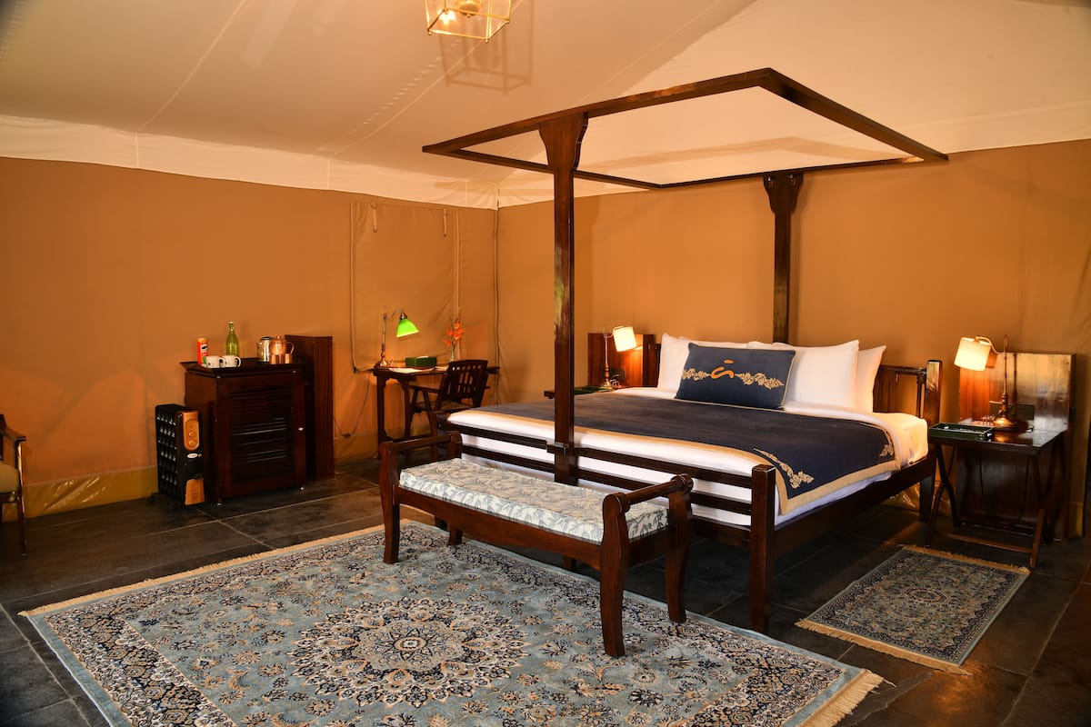 A four-poster bed with a bench, a nightstand, a desk, and a chair in a warmly lit room with patterned rugs.