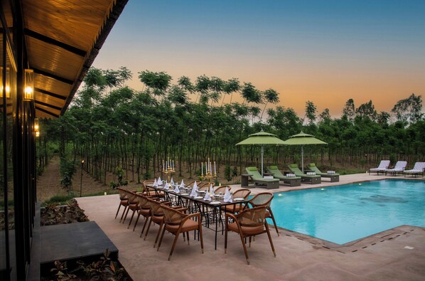 A poolside dining area with a table set for six, surrounded by lounge chairs and umbrellas, overlooking a pool with a forest backdrop.