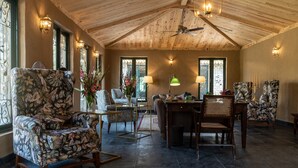 Een ruime woonkamer met een hoge plafond, houten balken en een plafondventilator. Er zijn meerdere zithoeken, waaronder een fauteuil met bloemenprint en een houten eettafel met stoelen.