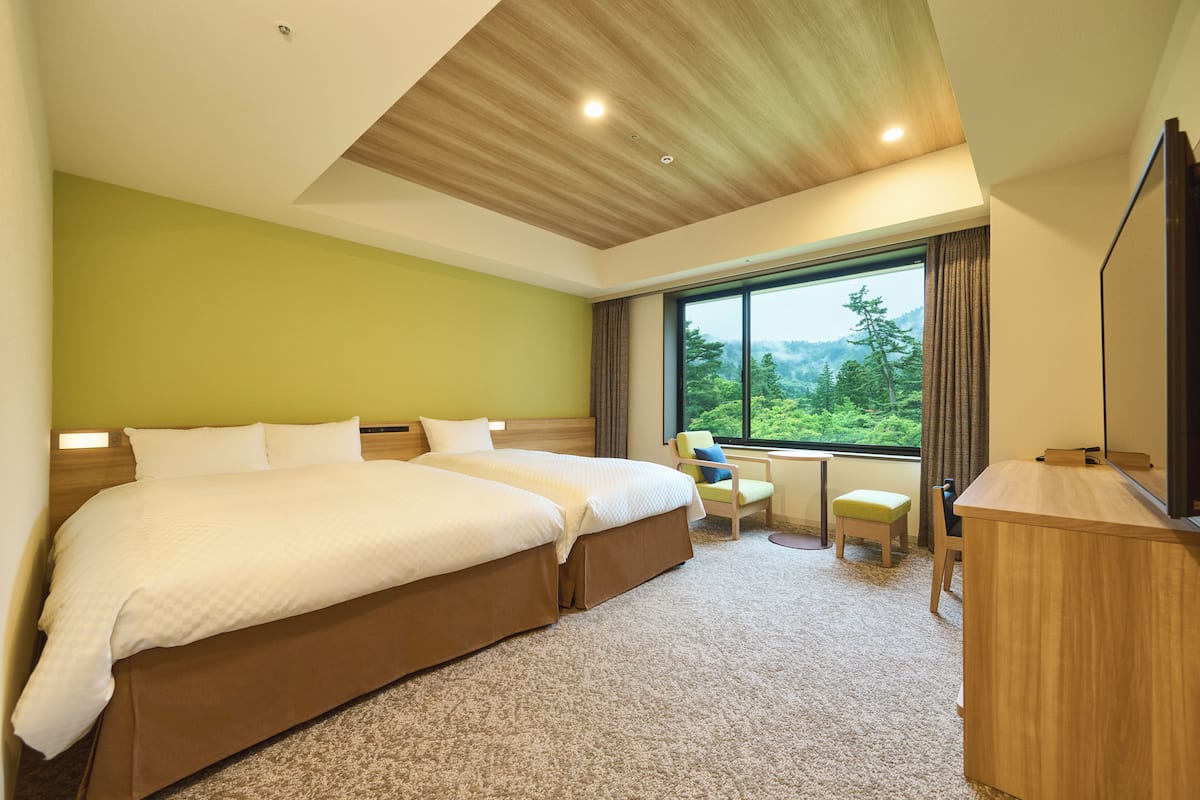 A hotel room with two beds, a desk, and a large window offering a view of greenery.