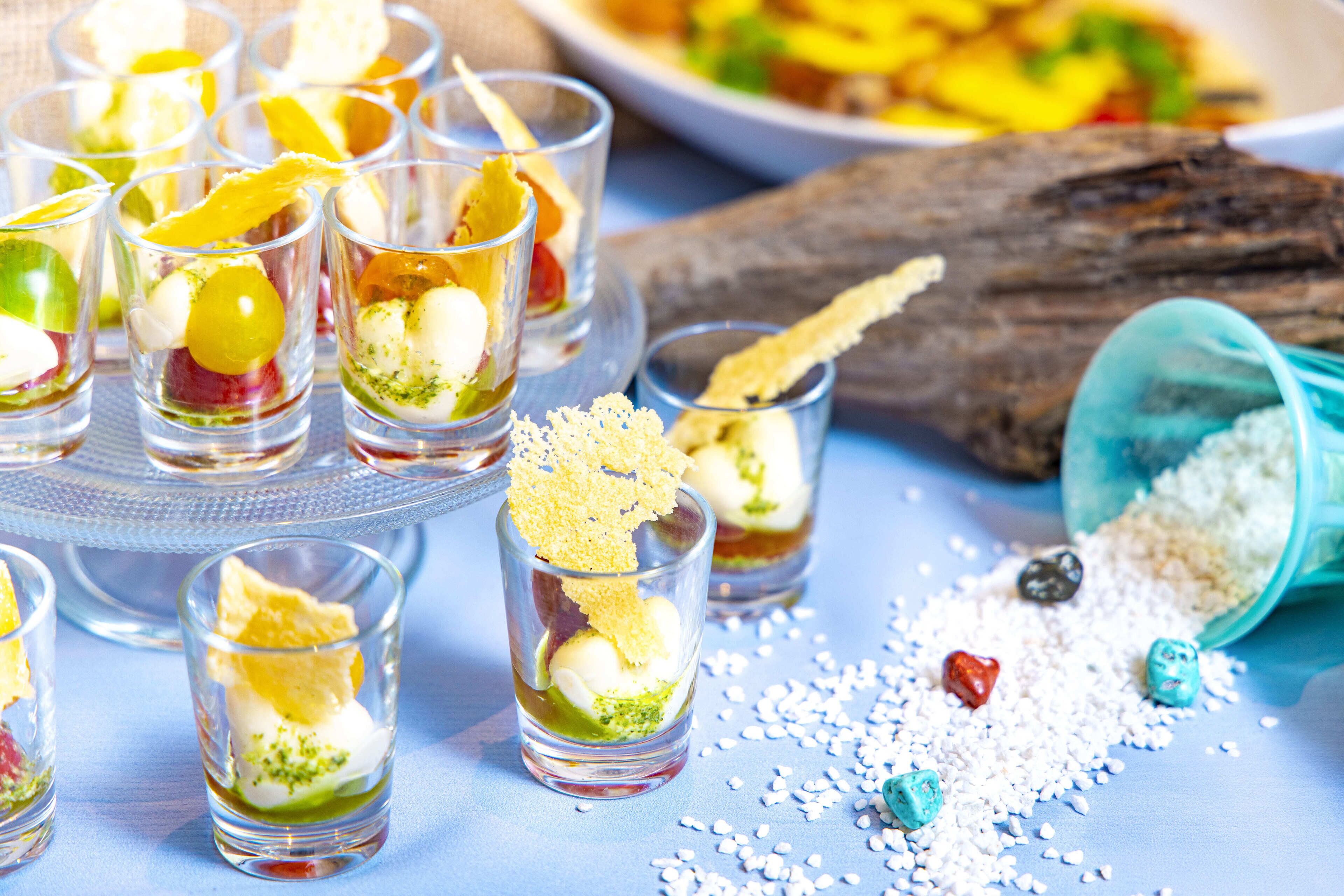 Une variété de petits verres à shooter remplis d’ingrédients colorés, disposés sur une surface bleue, avec une bûche en bois et une assiette de nourriture en arrière-plan.