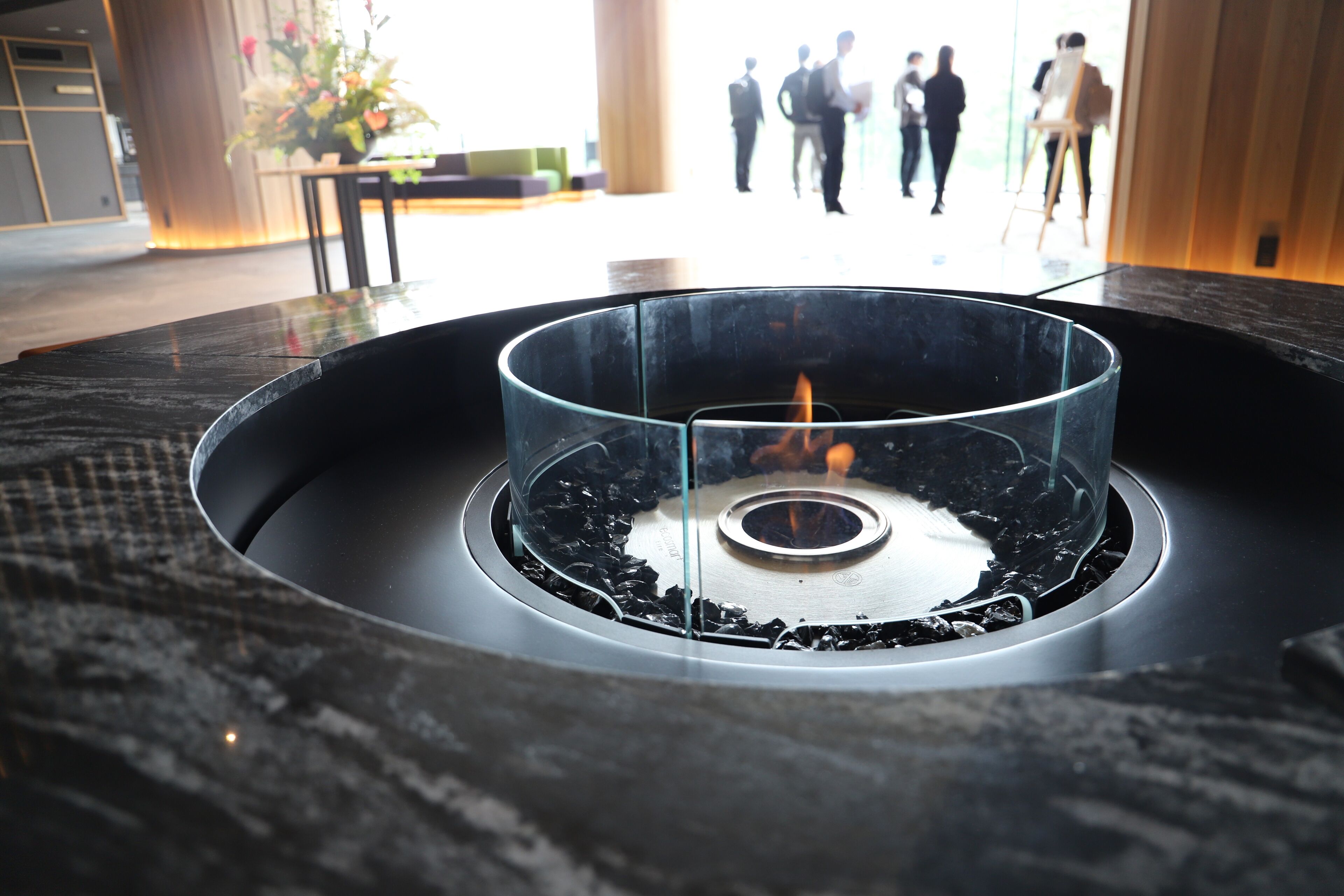 Un espacio interior moderno con una chimenea de vidrio central rodeada por un marco metálico circular.