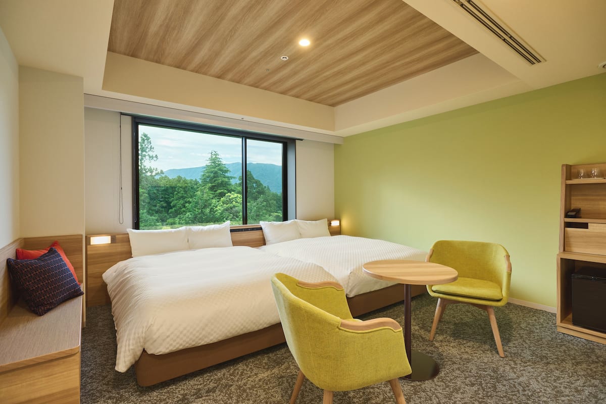 A modern hotel room with a large bed, a small round table, two chairs, and a view of greenery through the window.