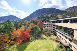 A modern building with a glass facade, surrounded by a lush garden and autumn-colored trees, set against a backdrop of mountains. - Hakone Kowakien (Hakone)