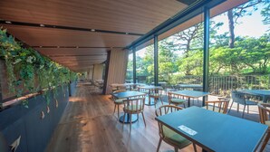 Un comedor interior con mesas y sillas de madera, ventanas amplias que ofrecen vistas a árboles, y una pared con vegetación.