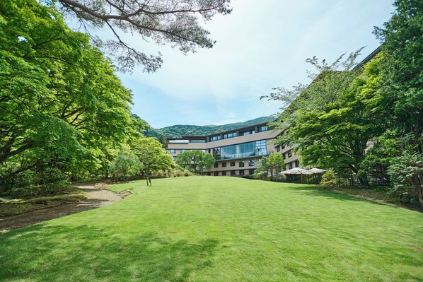 A courtyard with a grassy lawn, surrounded by trees and a modern building with multiple floors and large windows. - Hakone Kowakien (Hakone)