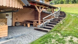 A wooden cabin with a stone-paved entrance, wooden benches, and a staircase leading to a covered area.