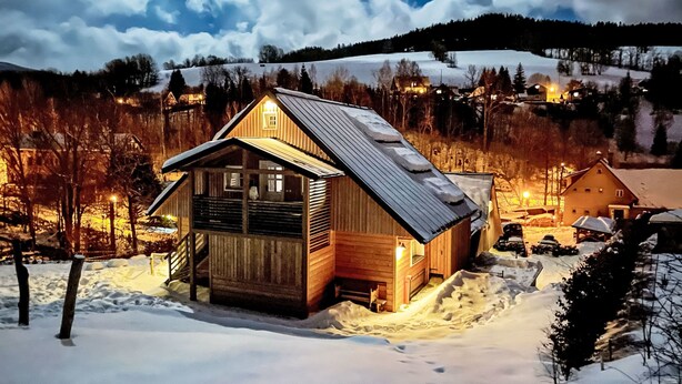 A wooden cabin with a sloped roof, surrounded by snow-covered ground and trees.