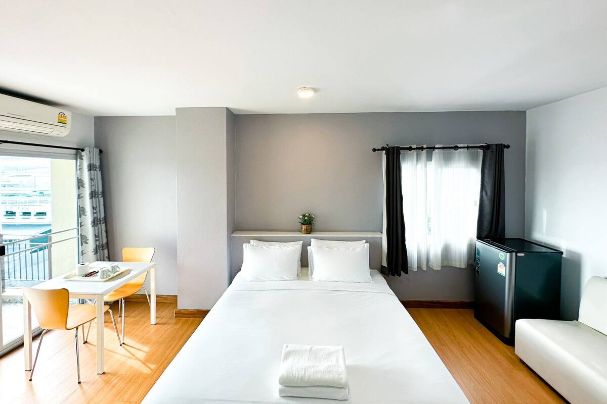 A hotel room with a bed, a desk, a chair, a small table, a refrigerator, and a window with curtains.