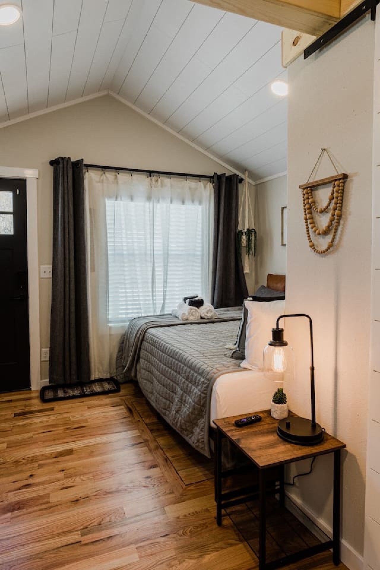 A bedroom with a bed, a bedside table with a lamp, a window with curtains, and a wooden floor.