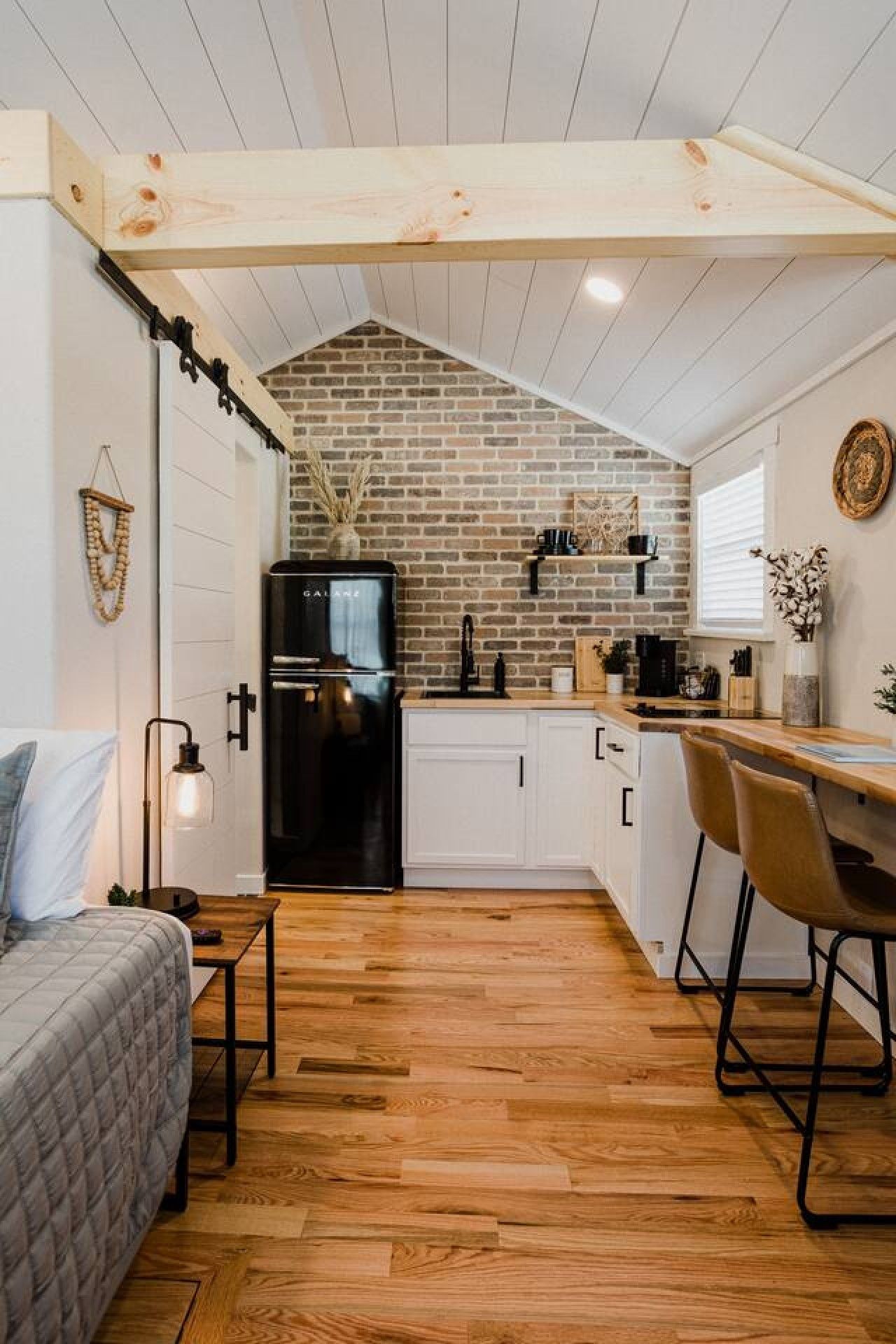 Cocina compacta con salpicadero de ladrillo, muebles blancos y refrigerador negro.