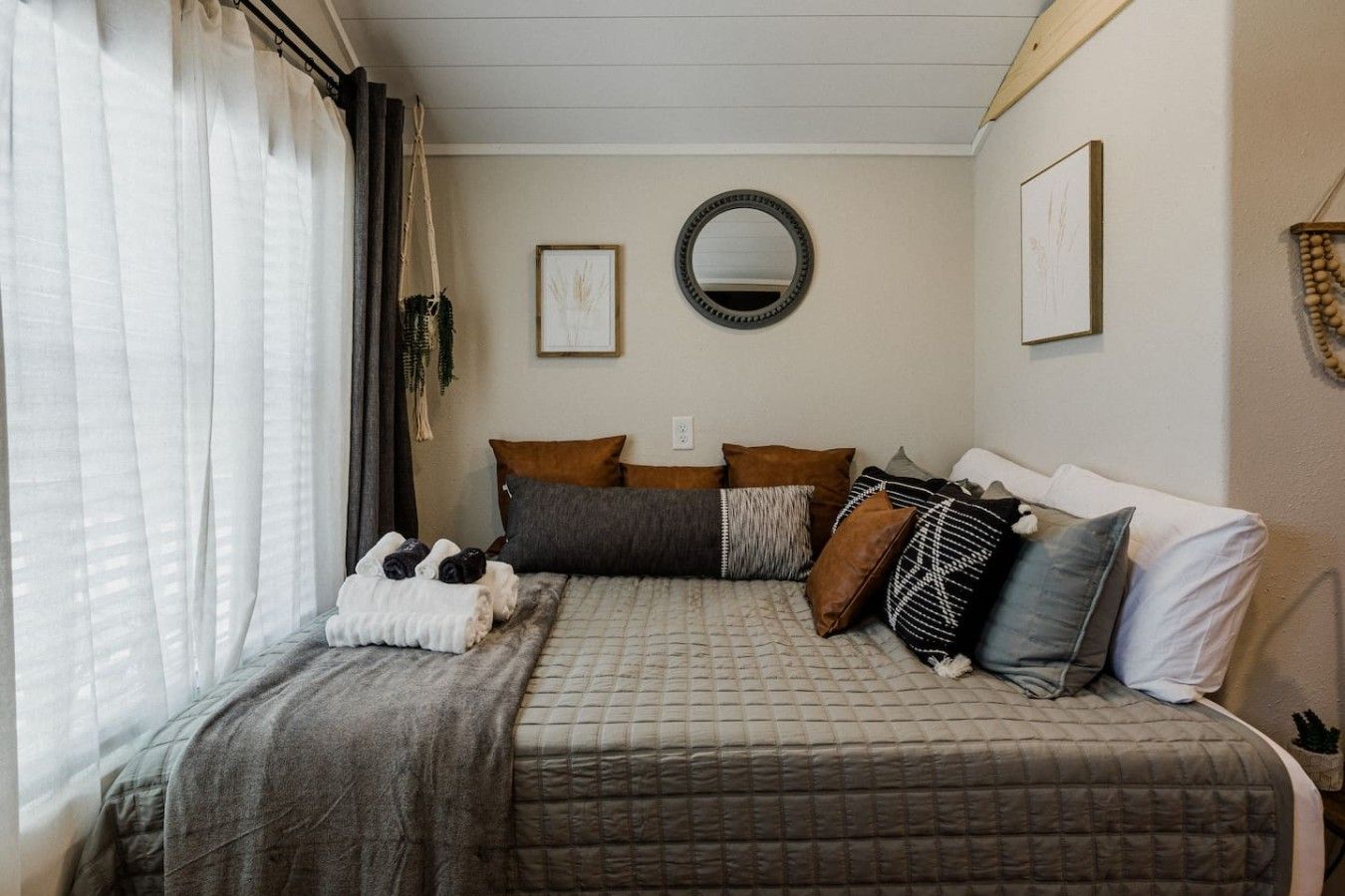 Une chambre à coucher avec un lit, des oreillers, une table de chevet, un miroir et une fenêtre avec des rideaux.