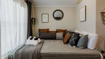 Une chambre à coucher avec un lit, des oreillers, une table de chevet, un miroir et une fenêtre avec des rideaux.