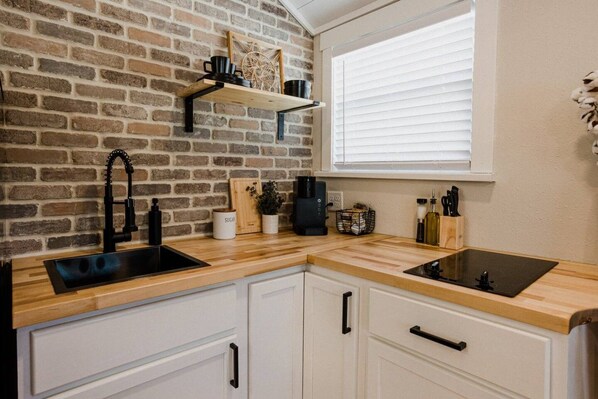 A kitchen with a brick wall, wooden countertop, white cabinets, a black faucet, and a window with blinds.