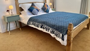 Una cama de madera con colcha azul y almohadas blancas, una mesita de noche con lámpara y una ventana con cortinas.