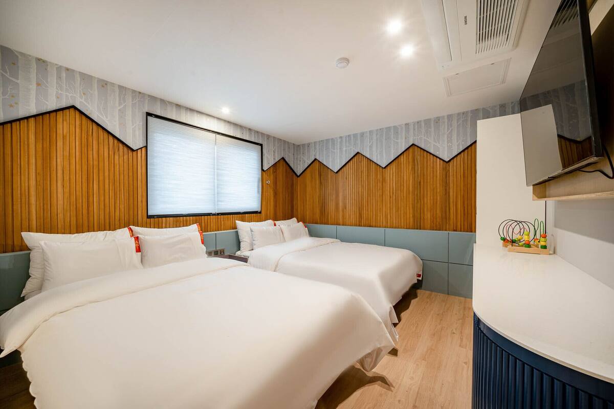 A hotel room with two beds, a wooden accent wall, a large window, and a built-in TV.