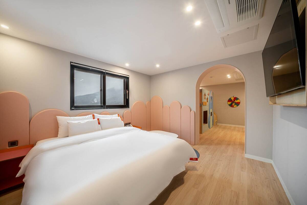 A modern hotel room with a large bed, a TV, a window, and a hallway leading to a dining area.