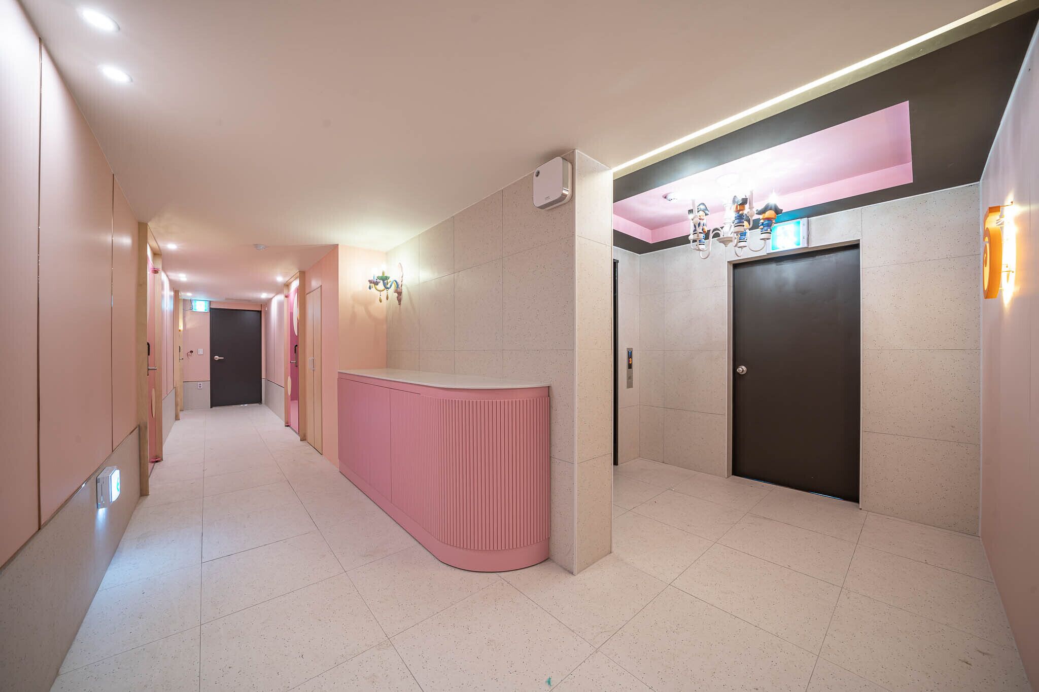 Un pasillo con un mostrador de recepción rosa, una puerta y una lámpara fijada a la pared.