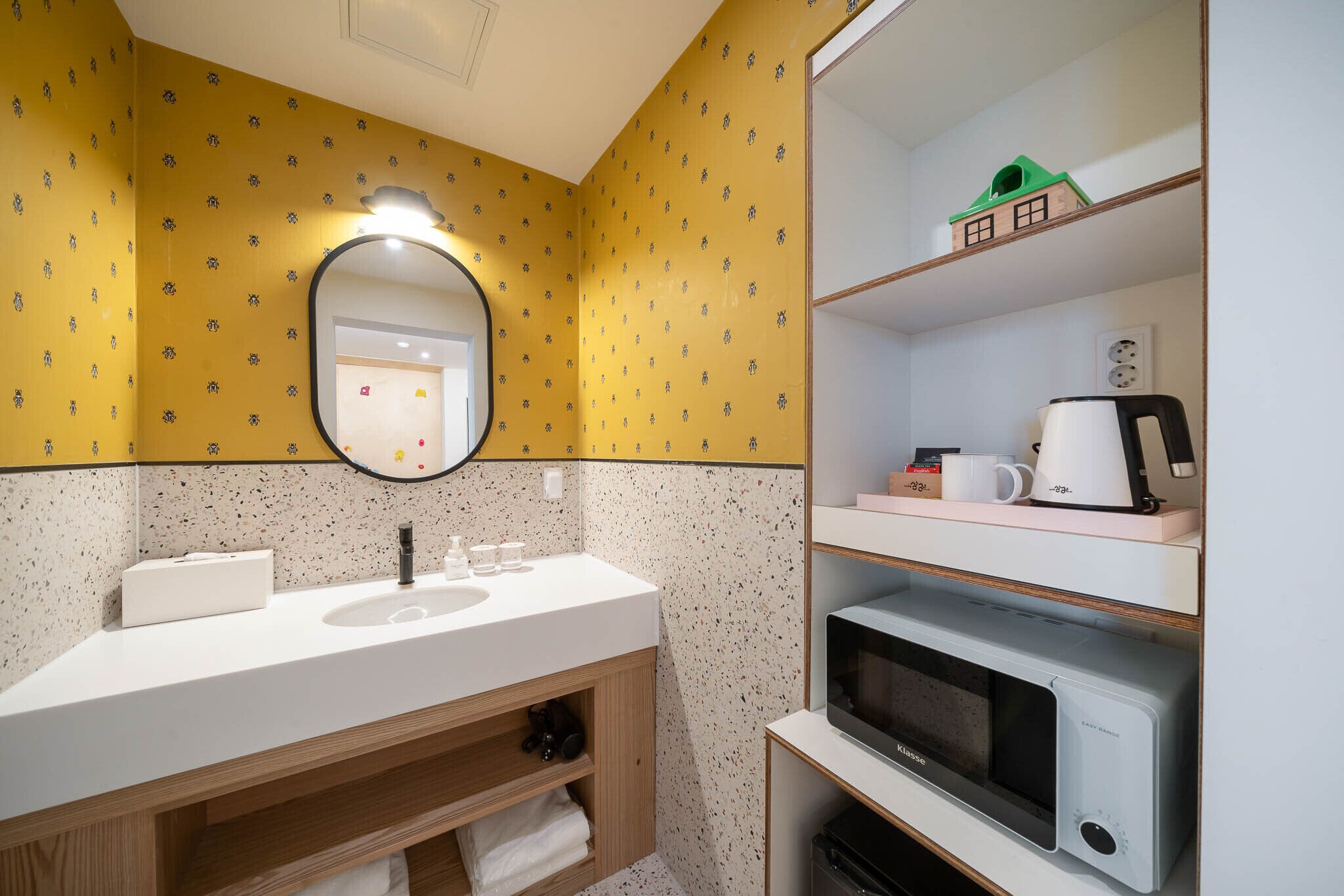 Baño con lavamanos blanco, espejo, repisa con modelo de casa verde, hervidor y microondas.