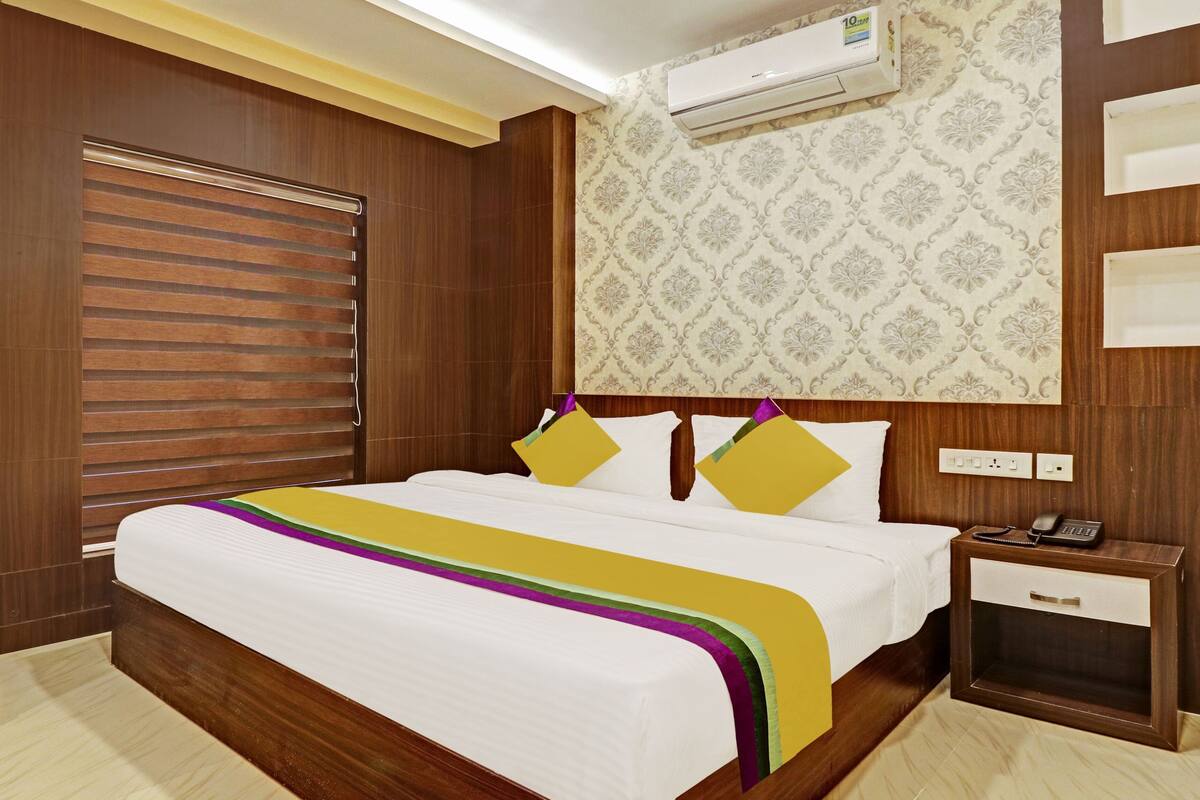 A hotel room with a wooden headboard, a patterned wallpaper, a bed with a white and colorful bedspread, and a bedside table with a phone and a lamp.