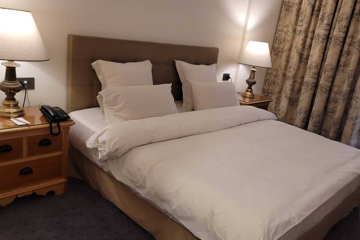 A hotel room with a bed, two bedside tables, a lamp, a phone, and a curtain.