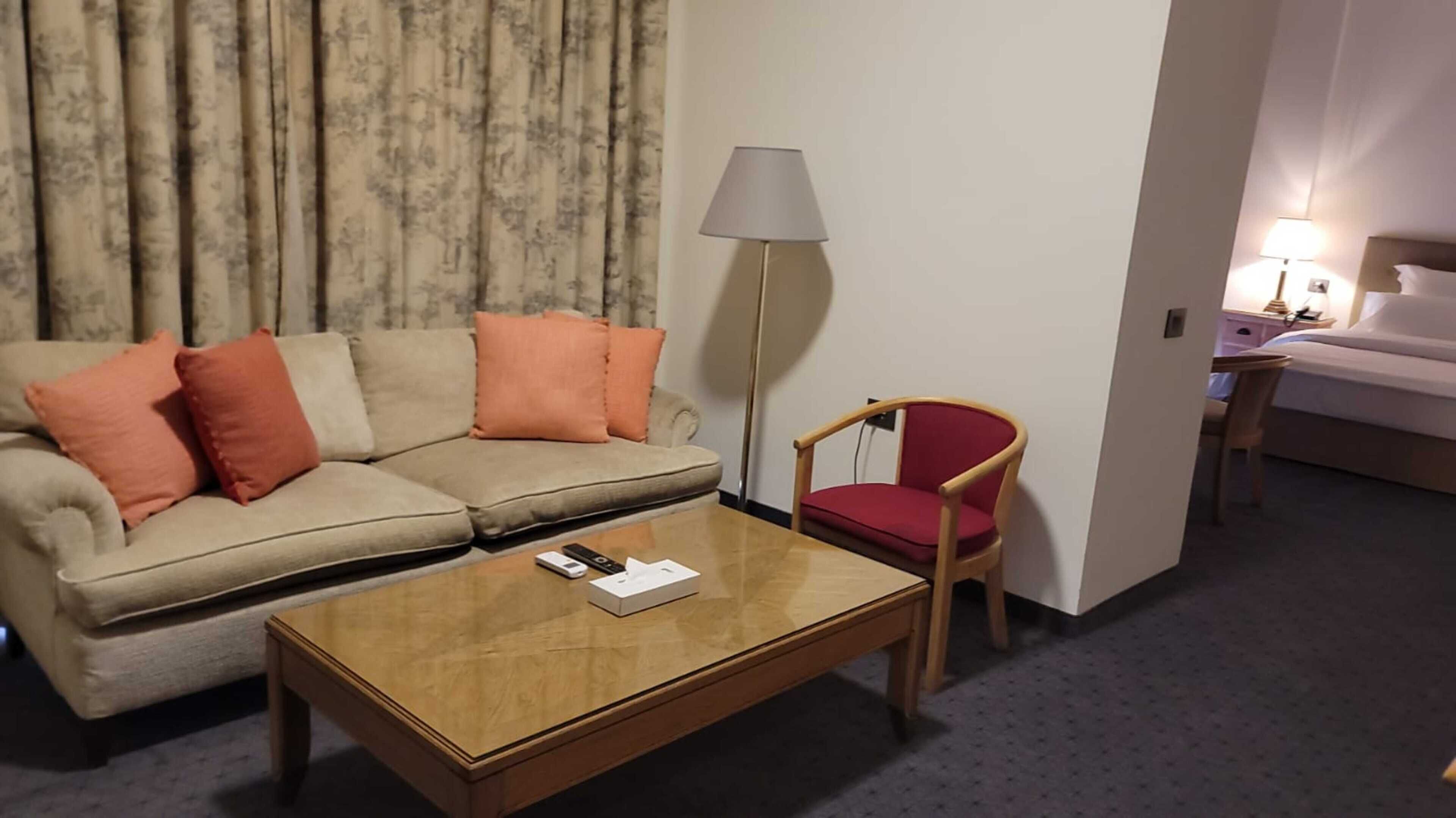 A hotel room with a sofa, a red chair, a wooden coffee table, a lamp, and a bed.