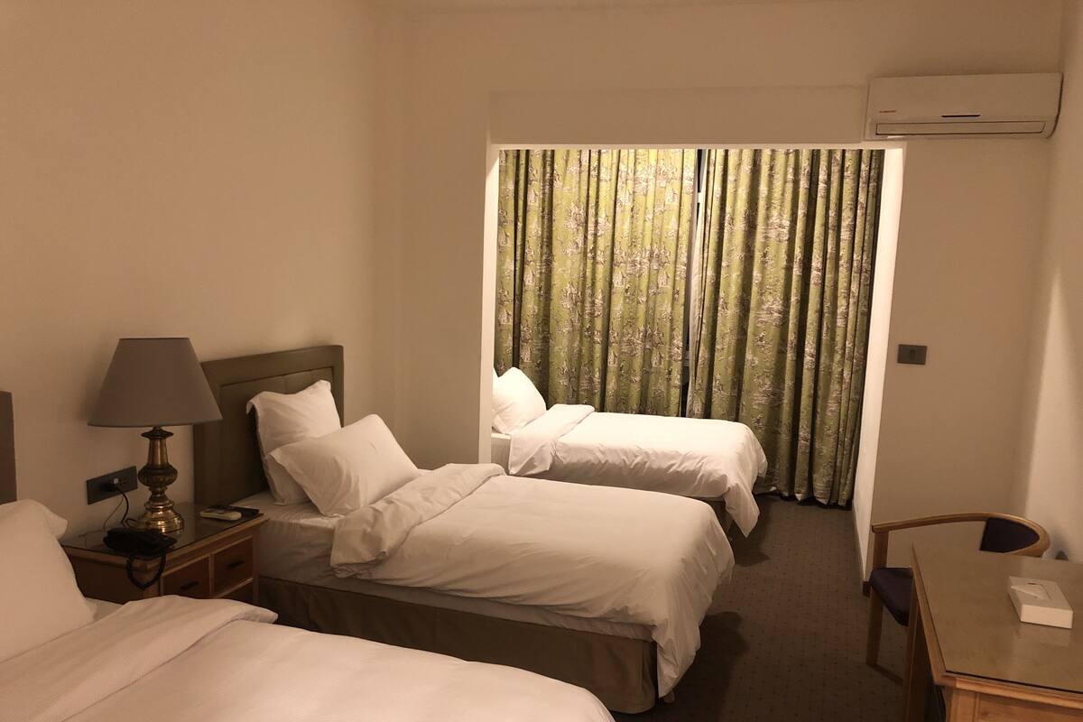 A hotel room with two beds, a bedside table with a lamp, a chair, and a desk.