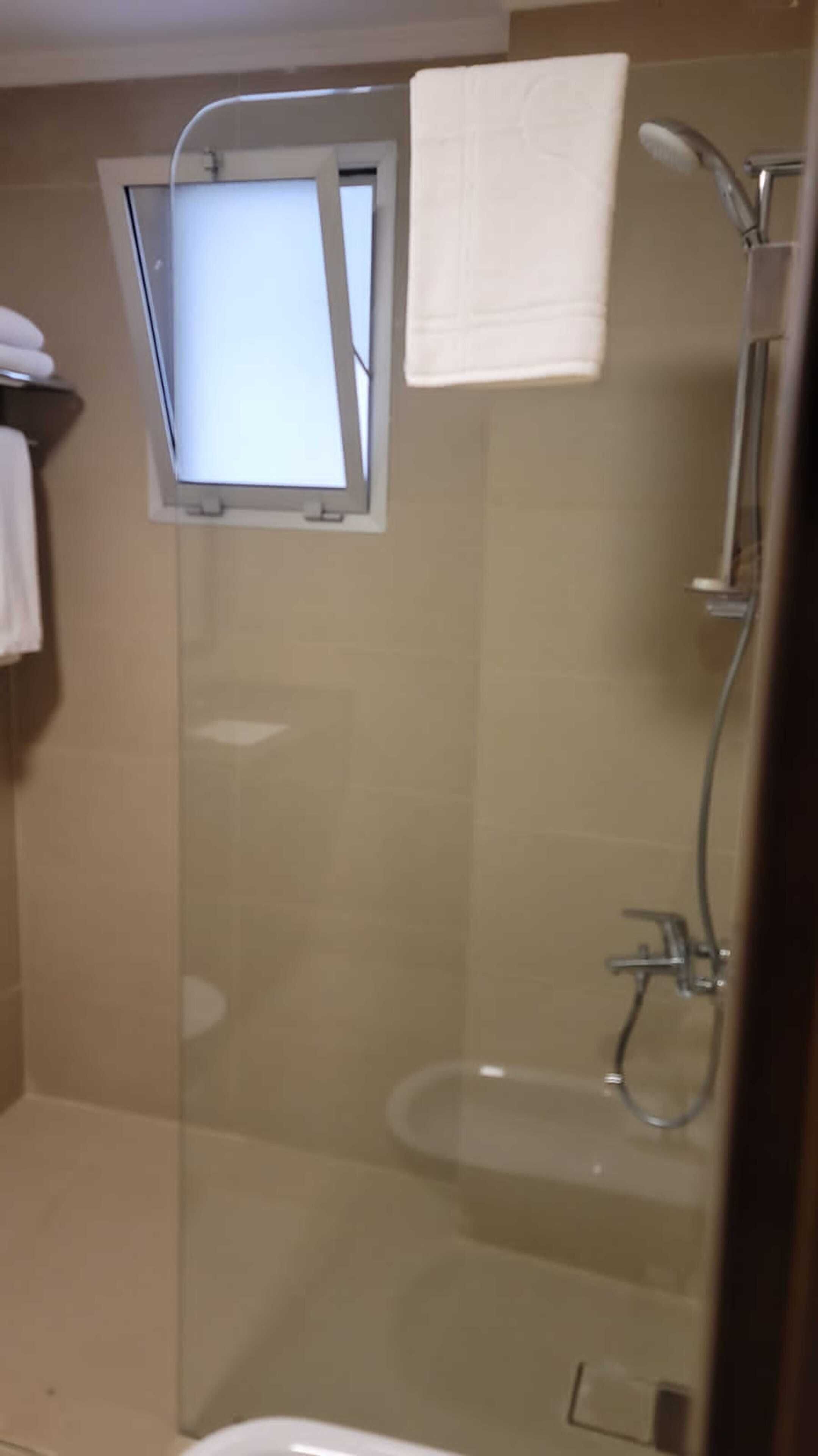 Ducha envuelta en vidrio con cabezal de ducha manual, ventana y una toalla colgada en la pared.