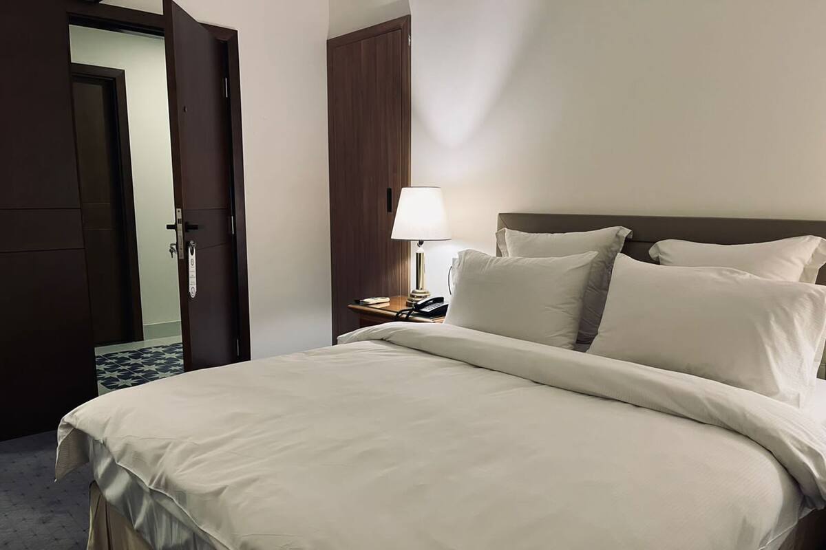 A hotel room with a large bed, a bedside table with a lamp, and a door leading to another room.