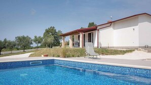 Ein Schwimmbecken mit gefliestem Rand, eine überdachte Terrasse und ein Gebäude mit rotem Dach und weißen Wänden.