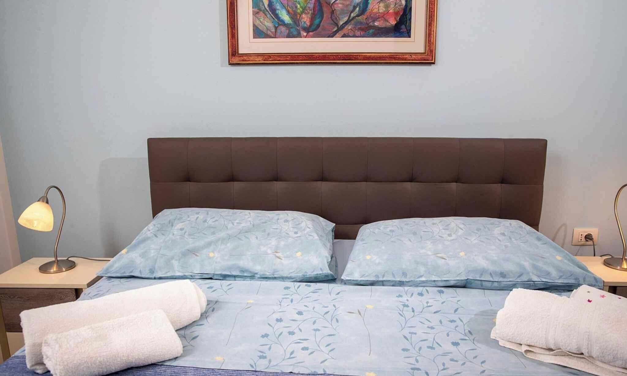 A bedroom with a brown upholstered headboard, two bedside tables with lamps, a floral-patterned bedspread, and two rolled towels on the bed.